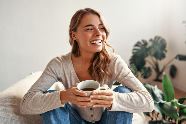 Young woman with a cup of hot coffee or tea sitting on a sofa in a cozy living room at home relaxing and unwinding on a weekend day.